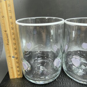May include: Two clear drinking glasses with a white rim and lavender heart-shaped designs. A wooden ruler with black markings is placed next to the glasses. The glasses are approximately 10 cm tall.