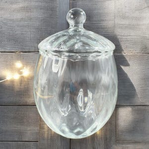 May include: A clear glass jar with a lid. The jar has a rounded, bulbous shape and a smooth, slightly textured surface. The lid has a knob on top.