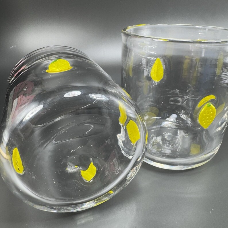May include: Two clear glass tumblers with yellow lemon designs. One tumbler is lying on its side, while the other stands upright. The lemons are painted on the outside of the glass.