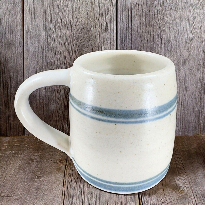 May include: Cream-colored ceramic mug with a rounded body and a curved handle. The mug features two horizontal blue stripes around the middle and bottom. The mug is set against a wooden background.