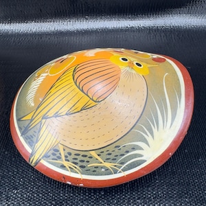 May include: Oval-shaped, hand-painted decorative dish featuring a stylized bird design. The bird has yellow, orange, and brown feathers, with a red border. The background has a gradient of colors, including yellow, green, and brown.