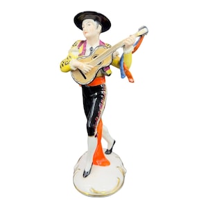 May include: A porcelain figurine of a man dressed in a traditional Spanish costume playing a guitar. He is wearing a black hat, a yellow and red vest, black pants, and red and white shoes. The figurine is standing on a white base.