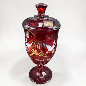 May include: A ruby red glass candy dish with a matching lid, featuring a gold-colored graphic of two white doves, a ribbon, and the text 40th Anniversary. A Viking brand sticker is visible on the lid. The vessel has a pedestal base.