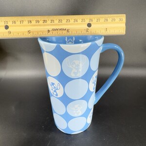 May include: A blue ceramic mug with a white polka dot pattern and a cartoon character of Stitch from Lilo & Stitch. The mug has a handle and is approximately 6 inches tall.