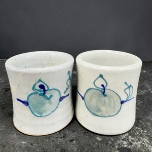 May include: Two handmade ceramic cups with a white speckled glaze. Each cup features a hand-painted design of a teal apple with blue accents. The cups are cylindrical with a slightly flared top, perfect for coffee or tea.