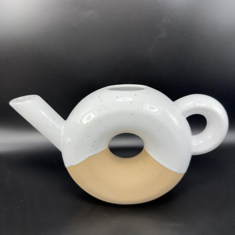 Peut inclure: Une th&eacute;i&egrave;re en c&eacute;ramique blanche et beige en forme de donut. La th&eacute;i&egrave;re a un bec verseur, une anse et un trou central. La moiti&eacute; sup&eacute;rieure est blanche avec des mouchetures noires, et la moiti&eacute; inf&eacute;rieure est beige.