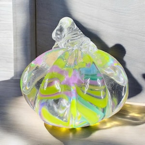 May include: A clear glass decorative pumpkin with a swirled pattern of yellow, green, purple, and blue. The pumpkin has a textured surface and a clear, twisted stem.