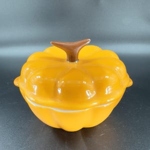 May include: A bright orange ceramic lidded casserole dish shaped like a pumpkin. The lid features a brown stem-shaped handle and a ribbed, segmented surface that mimics the appearance of a gourd. The base is rounded with matching ribs.