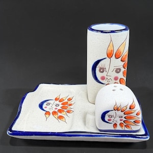 May include: A ceramic salt and pepper set with a matching tray. The set features a white background with a blue trim and is decorated with a sun and moon design in orange, blue, and pink. The set includes a salt shaker, pepper shaker, and a utensil holder.