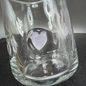 May include: A clear glass vase with a heart-shaped design. The vase has a textured surface and a rounded shape. The heart-shaped design is in the center of the vase and is filled with a light purple colour.
