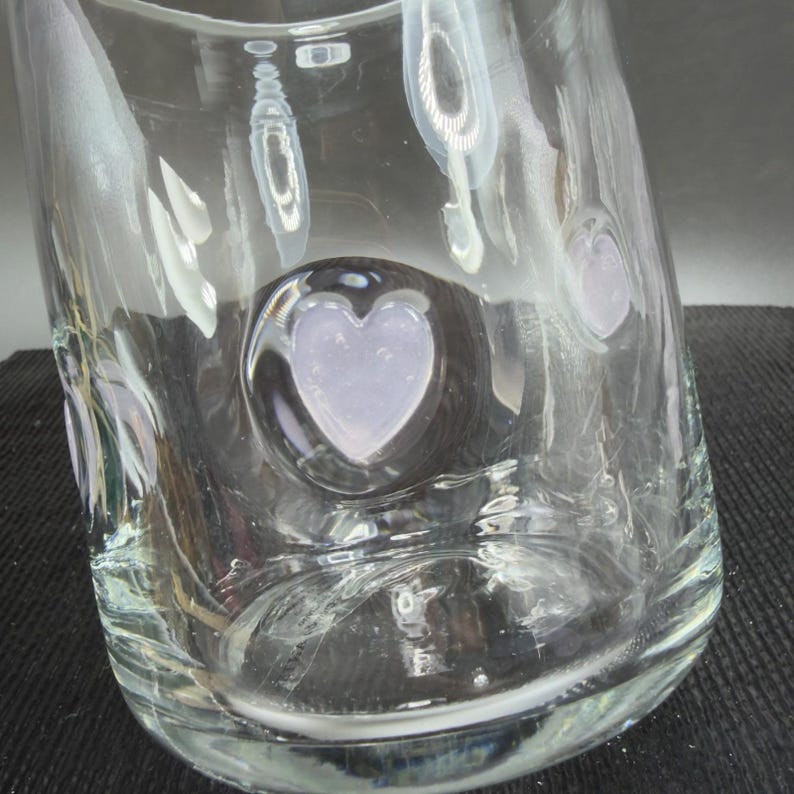 May include: A clear glass vase with a heart-shaped design. The vase has a textured surface and a rounded shape. The heart-shaped design is in the center of the vase and is filled with a light purple colour.
