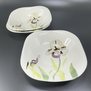 May include: Set of three off-white ceramic dishes. Each dish features a hand-painted iris flower design in shades of green, purple, and yellow. The dishes have a square shape with rounded edges, perfect for serving appetizers or desserts.