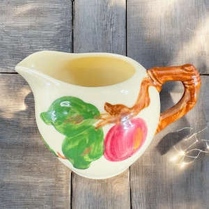 May include: A vintage cream-colored ceramic creamer with a brown handle. The creamer features a painted design of a red apple with green leaves on a branch.
