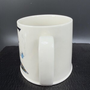 May include: White ceramic mug with a curved handle. The mug features a design with blue, yellow, and black accents. The mug is cylindrical with a rounded base.