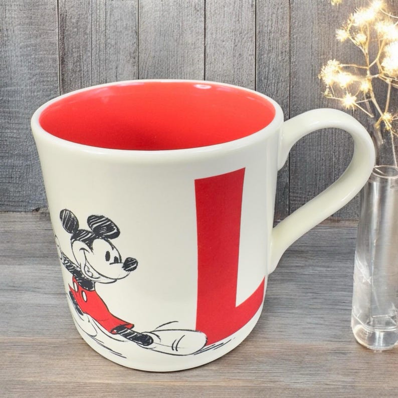 May include: Cream-colored ceramic mug with a red interior and a large red letter "L". The mug features a smiling Mickey Mouse graphic in black and red. The handle is curved and the mug sits on a wooden surface.