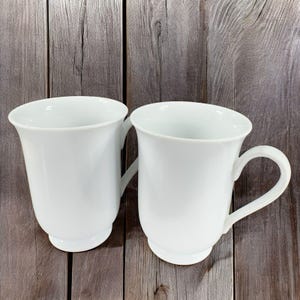 May include: Two white ceramic mugs with a rounded shape and a handle. The mugs are sitting on a wooden surface.