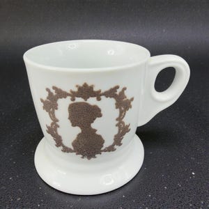 May include: White ceramic mug with a brown silhouette of a woman in a decorative frame. The mug has a handle and a wide base. The design is a classic, vintage style.