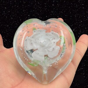 May include: A heart-shaped glass paperweight with a white flower and green leaves inside. The glass is filled with small air bubbles.