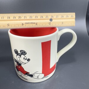 May include: White ceramic mug with a red interior and a large red letter "L". The mug features a black and red illustration of Mickey Mouse. The mug is approximately 4 inches tall.