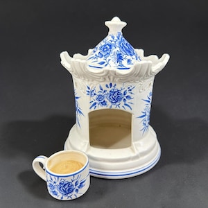 May include: A white ceramic decorative pagoda-shaped structure with blue floral designs and a small matching cup. The cup contains a light brown liquid. The pagoda has a domed top and open sides.