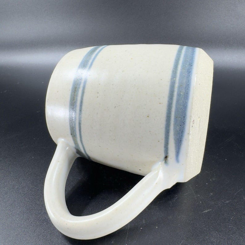 May include: Cream-colored ceramic mug with two vertical blue stripes. The mug has a rounded handle and a speckled texture. The stripes are slightly uneven, adding to the handmade aesthetic. The mug is suitable for hot or cold beverages.