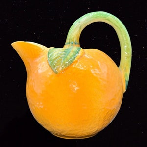 May include: A ceramic pitcher shaped like an orange with a green leaf and stem. The pitcher is glazed in a bright orange color and has a textured surface.