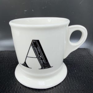 May include: White ceramic mug with a black letter "A" on the front. The mug has a rounded base and a handle. The letter "A" is in a bold, decorative font. The mug is likely for coffee or tea.