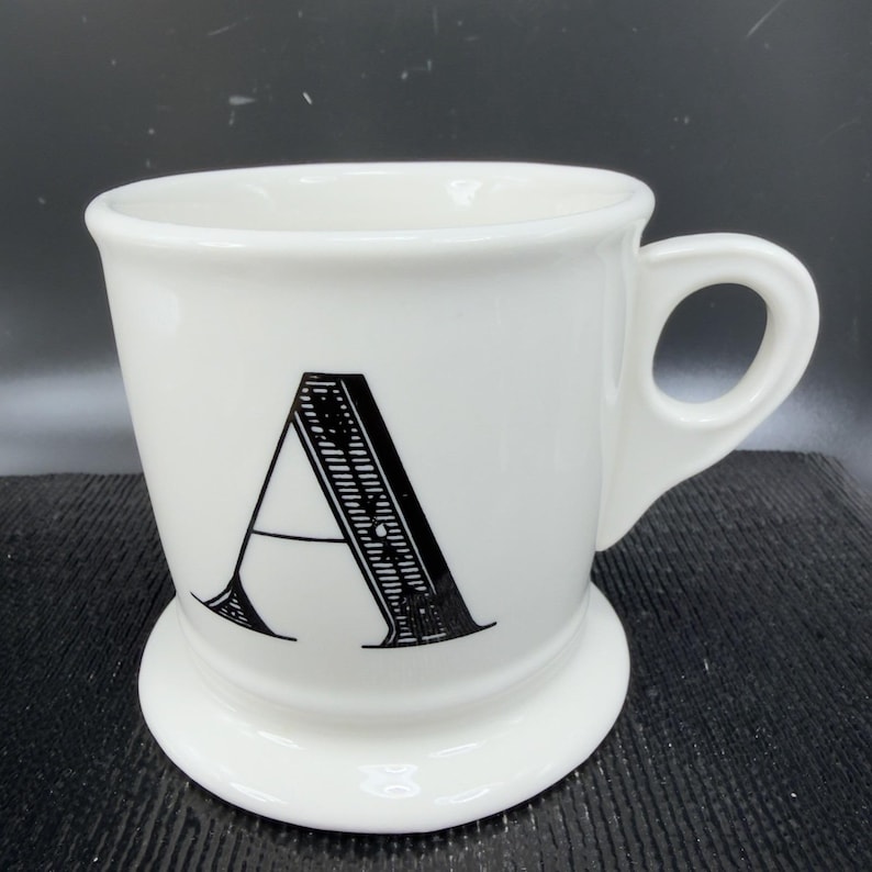 May include: White ceramic mug with a black letter "A" on the front. The mug has a rounded base and a handle. The letter "A" is in a bold, decorative font. The mug is likely for coffee or tea.