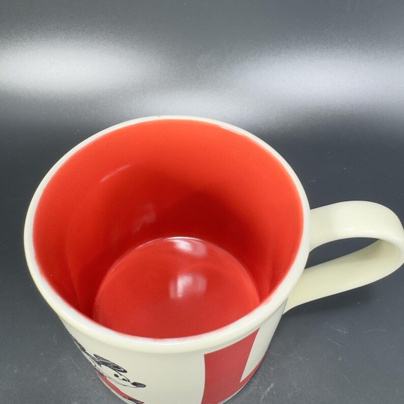 May include: A white ceramic mug with a red interior. The mug has a curved handle and a design on the exterior. The mug is empty and appears to be clean. The mug is likely used for drinking beverages.
