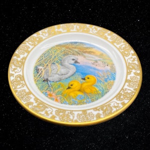 May include: A white porcelain plate with a gold rim and a floral design. The center of the plate features a painting of a swan and two ducklings in a pond setting.