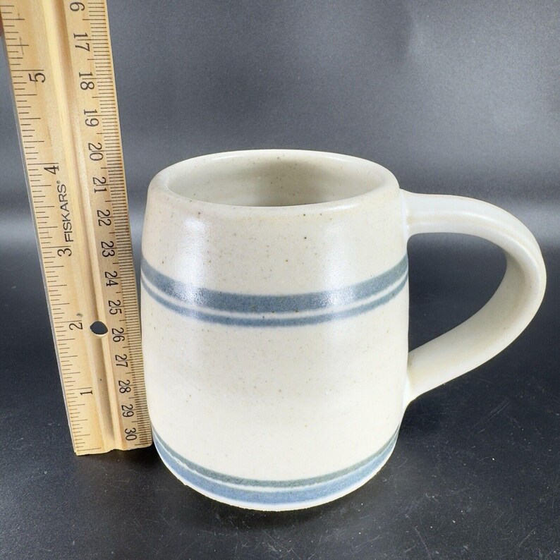 May include: Cream-colored ceramic mug with two horizontal blue stripes. The mug has a rounded body, a curved handle, and is approximately 4 inches tall. The mug is stoneware and has a speckled texture.