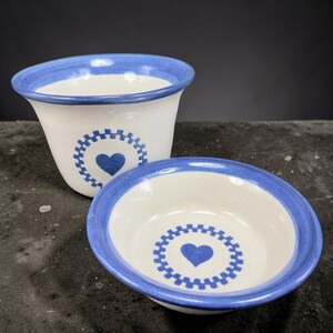 May include: Two ceramic bowls with a white base and blue trim. Each bowl features a blue heart in the center, surrounded by a decorative blue border. One bowl is a small, shallow dish, and the other is a taller, flared bowl.