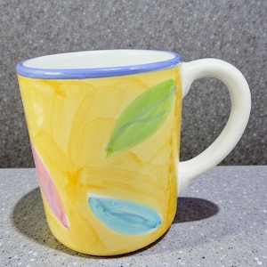 May include: Yellow ceramic mug with a white handle and a blue rim. The mug is decorated with colorful leaf-shaped designs in green, pink, and blue. The background has a yellow brushstroke pattern.