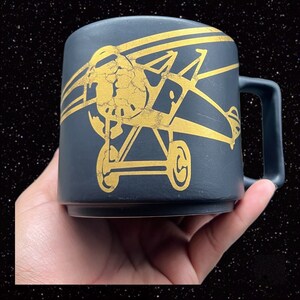 May include: Black ceramic mug with a gold-colored airplane design. The airplane is a biplane with a propeller and wheels. The mug has a handle.
