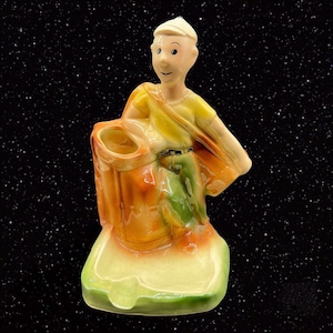 May include: A ceramic figurine of a golfer carrying a golf bag. The figurine is painted in shades of yellow, green, orange, and brown. The golfer is wearing a yellow shirt, green pants, and a brown hat. The golf bag is brown with a yellow top. The figurine is standing on a base that is painted in shades of green and yellow.