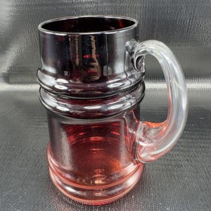 May include: A ruby red glass mug with a clear handle. The mug has a cylindrical body with a ridged design around the middle. The handle is arched and attached to the side of the mug. The glass is thick and appears to be hand-blown.