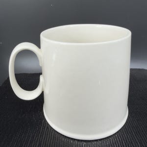May include: A white ceramic mug with a rounded handle. The mug has a cylindrical shape and a slightly flared base. The interior of the mug is also white. The mug is set against a dark background.