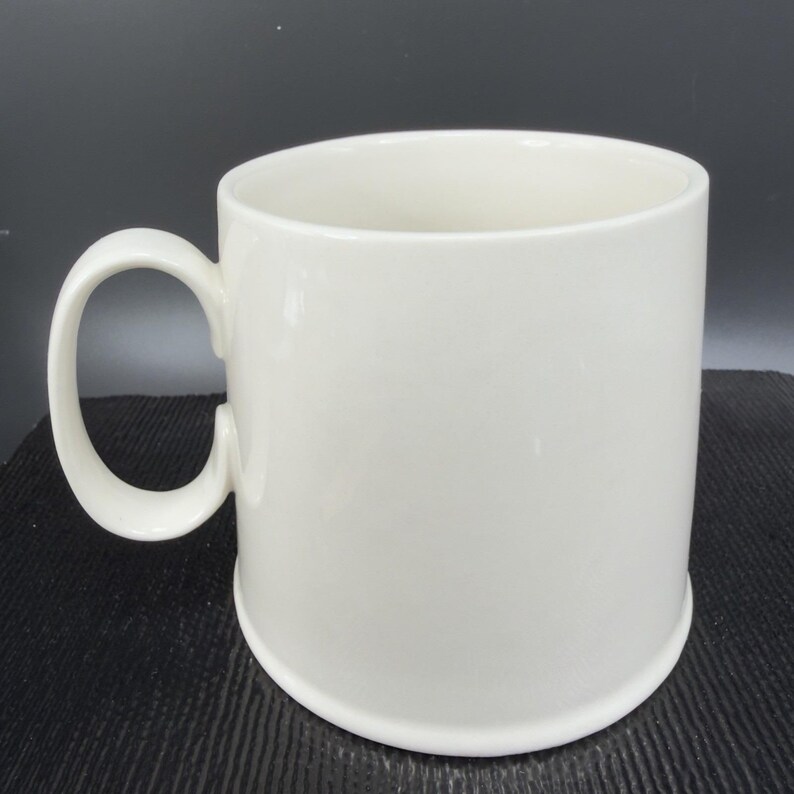 May include: A white ceramic mug with a rounded handle. The mug has a cylindrical shape and a slightly flared base. The interior of the mug is also white. The mug is set against a dark background.