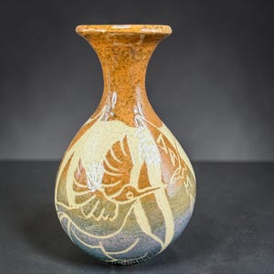 May include: A ceramic vase with a flared rim and a bulbous body featuring a gradient color scheme transitioning from orange at the top to cream and blue at the base. The surface is decorated with an etched line drawing of a hummingbird in flight.