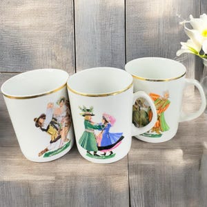 May include: Three white ceramic mugs with gold trim and colorful illustrations of people in traditional clothing dancing. The mugs are on a wooden surface.
