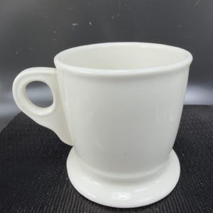 May include: A white ceramic mug with a handle and a pedestal base. The mug has a smooth, glossy finish and a simple, classic design. The mug is empty and ready to be filled with a beverage.