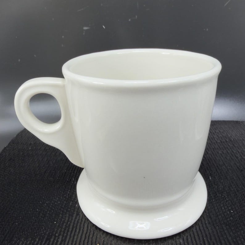 May include: A white ceramic mug with a handle and a pedestal base. The mug has a smooth, glossy finish and a simple, classic design. The mug is empty and ready to be filled with a beverage.