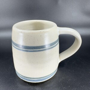 May include: A cream-colored ceramic mug with two horizontal blue stripes. The mug has a rounded body, a curved handle, and a speckled texture. The mug is suitable for coffee, tea, or other beverages.