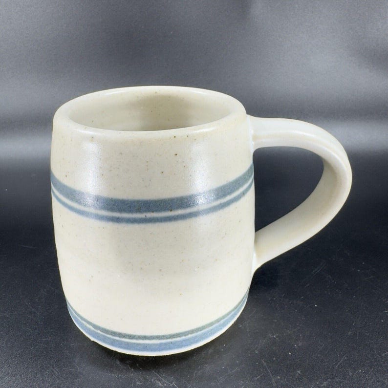 May include: A cream-colored ceramic mug with two horizontal blue stripes. The mug has a rounded body, a curved handle, and a speckled texture. The mug is suitable for coffee, tea, or other beverages.