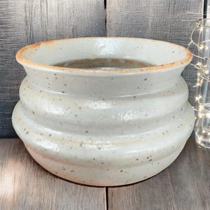May include: A handcrafted, off-white ceramic planter with a speckled finish. The planter features a wide, open top and a series of horizontal ridges around the body. The rim has a subtle, warm brown hue.