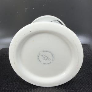 May include: White ceramic vase with a wide base and a bird logo on the bottom. The vase has a cylindrical shape and a slightly flared rim. The bottom of the vase has a circular stamp with text and a bird illustration.