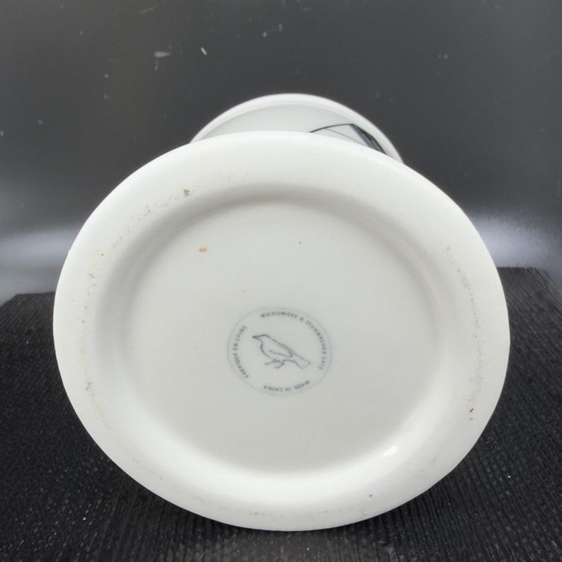May include: White ceramic vase with a wide base and a bird logo on the bottom. The vase has a cylindrical shape and a slightly flared rim. The bottom of the vase has a circular stamp with text and a bird illustration.