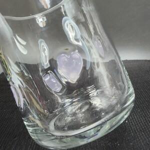 May include: A clear glass vase with a wide base and curved sides. The vase is decorated with white and lavender heart and oval shapes. The vase is sitting on a dark surface.
