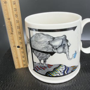 May include: White ceramic mug featuring a whimsical illustration of an elephant with a trunk holding a watering can. The elephant is standing on a colorful, patterned base. The mug has a black border and a curved handle.