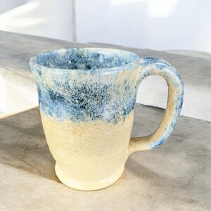 May include: A handmade ceramic mug with a blue and cream glaze. The mug has a rounded body and a handle.